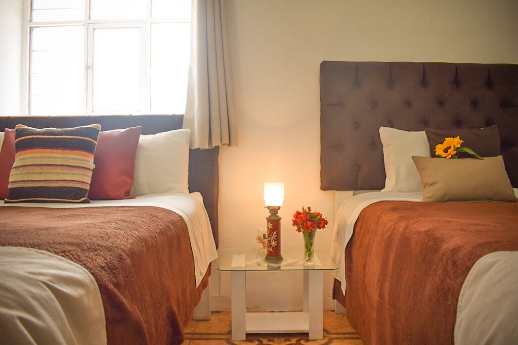 Ein Hotelzimmer mit zwei Betten, einem Nachttisch mit Lampe und Blumen sowie einem Fenster mit Vorhängen.
