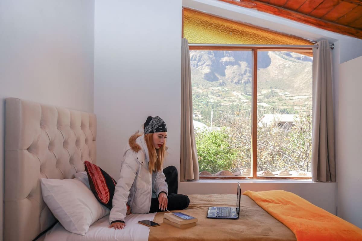 A room with a bed, a laptop, a book, and a window with a view of a mountainous landscape.