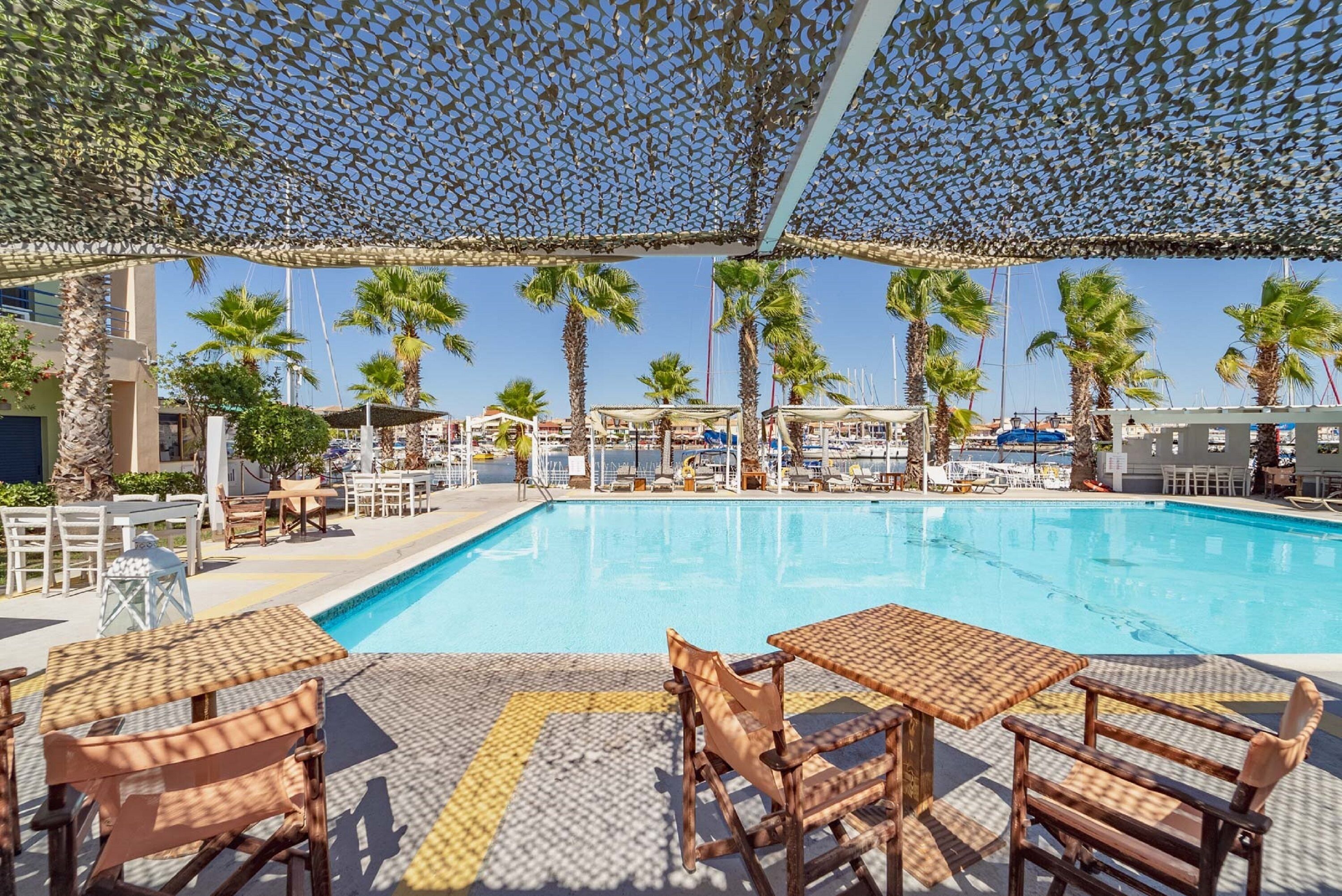 Un área de piscina con sillones de descanso y mesas, palmeras y un cielo azul despejado.