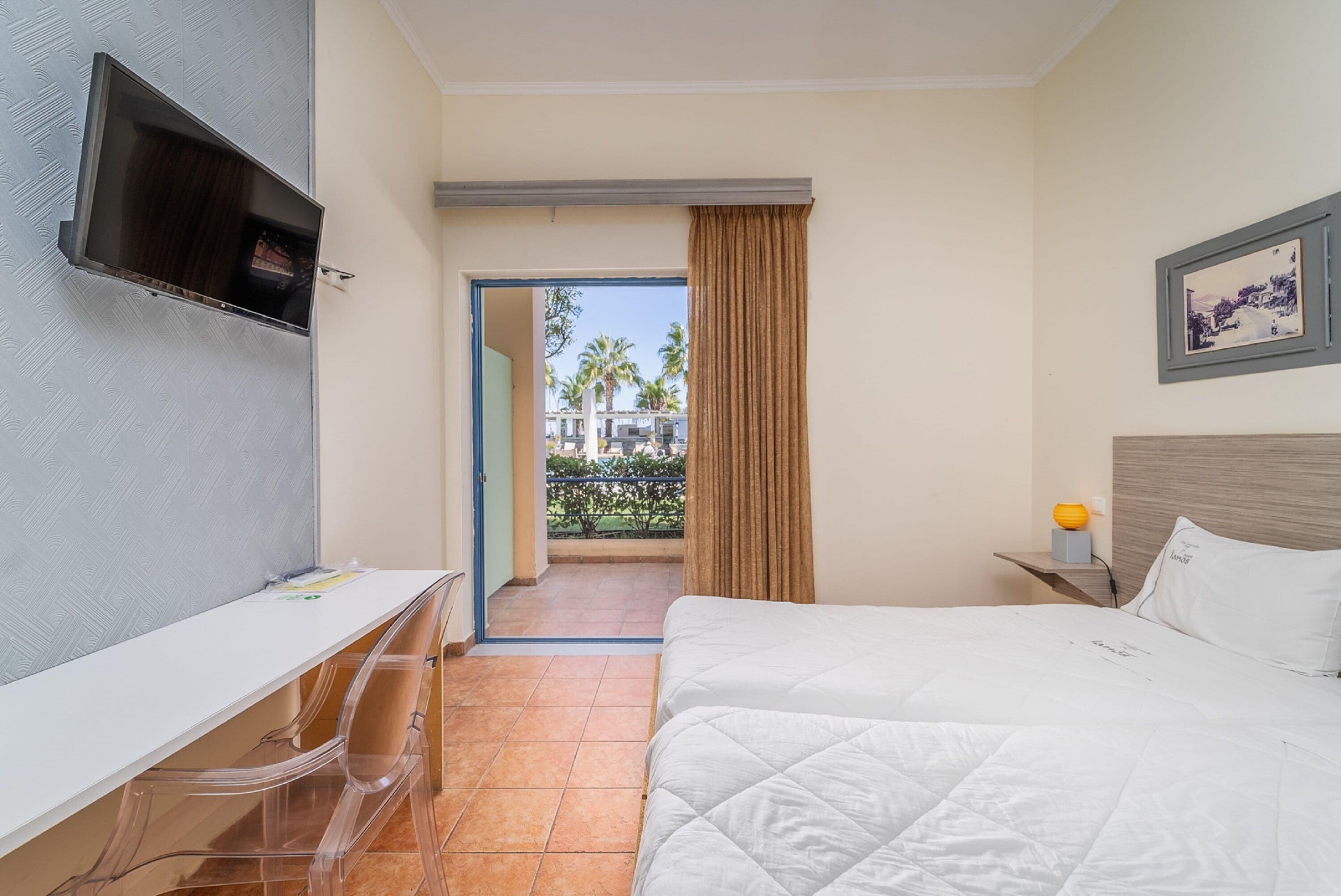 A hotel room with a bed, a desk, a TV, and a balcony with a view of palm trees.