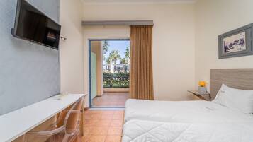 A hotel room with a bed, a desk, a TV, and a balcony with a view of palm trees.