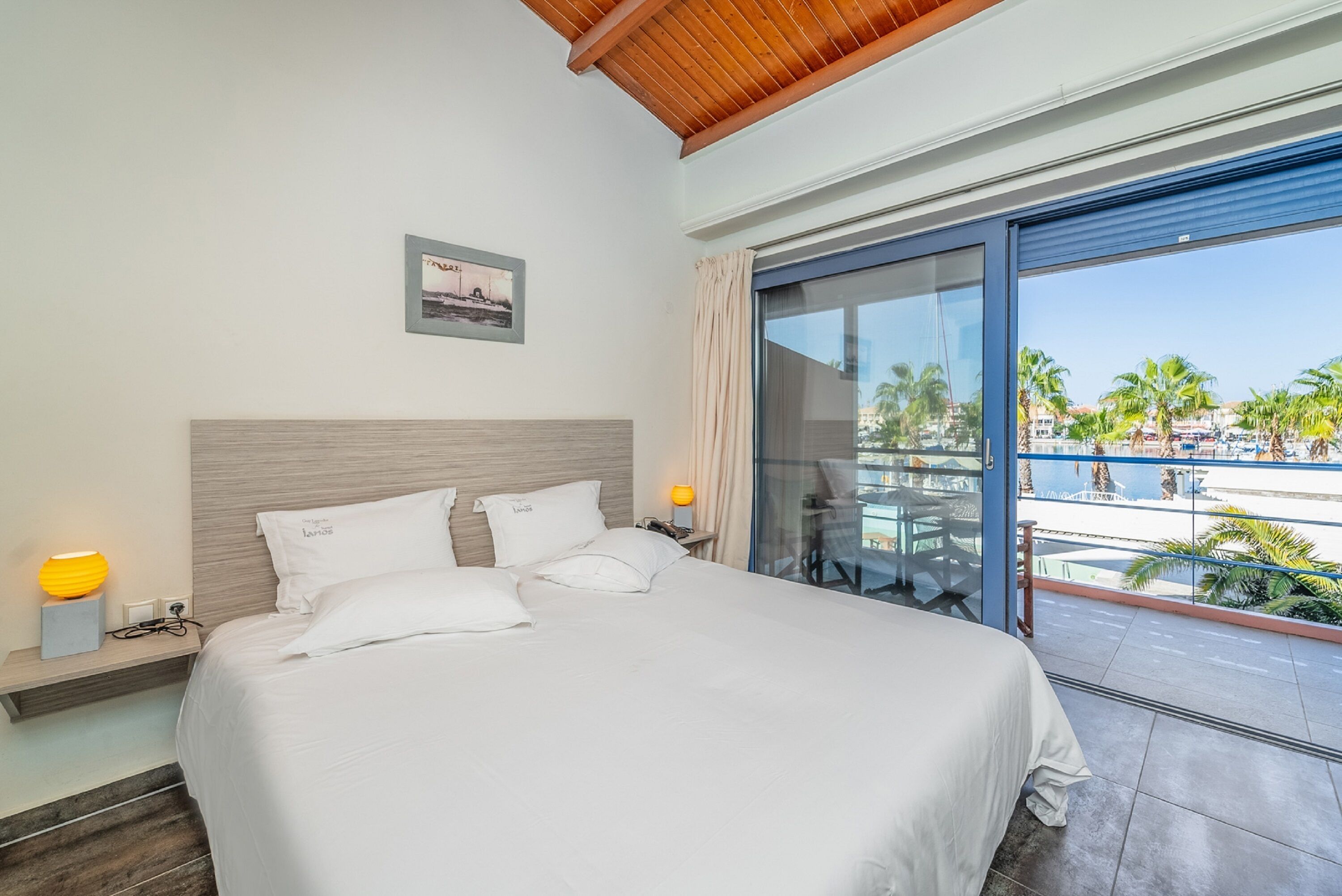 A hotel room with a large bed, a wooden ceiling, a balcony with a view of palm trees and a pool, and a framed picture on the wall.