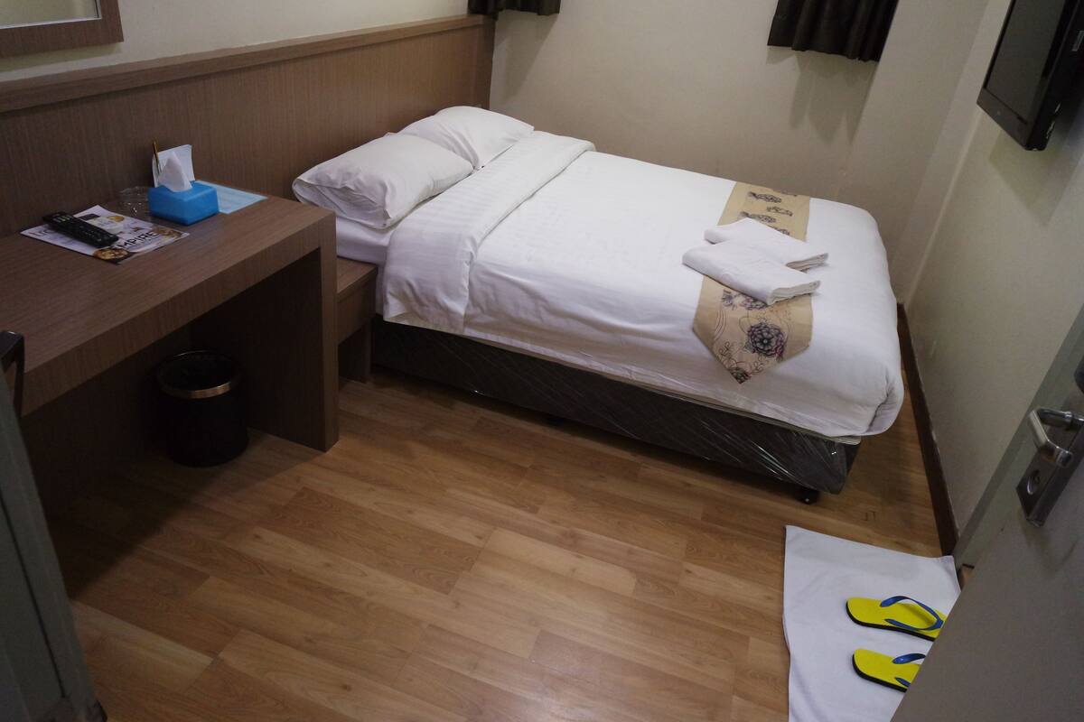 A small hotel room with a single bed, a wooden nightstand, a trash bin, and a pair of flip-flops on the floor.