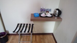A small kitchenette with a wooden shelf holding a kettle, two water bottles, a tray with cups, and a blue container. A metal drying rack is on the floor.