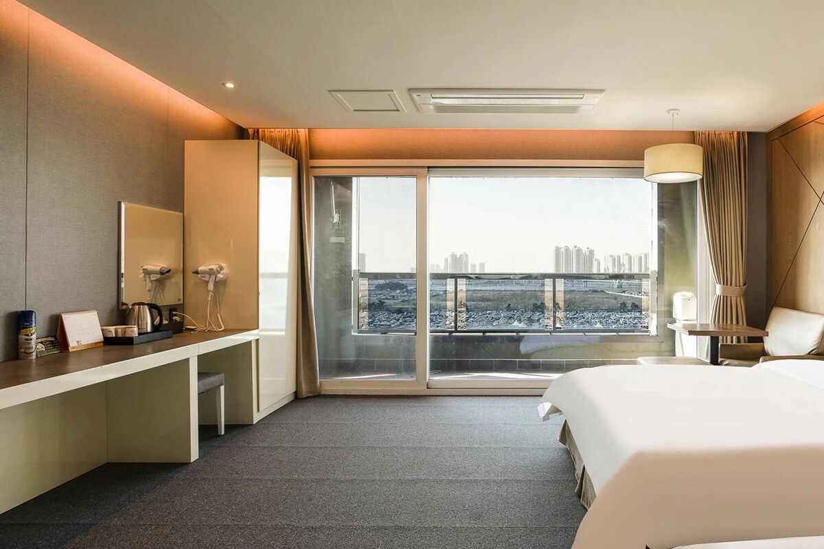 Une chambre d’hôtel avec un lit, un bureau, une chaise, une grande fenêtre et une vue sur le paysage urbain.
