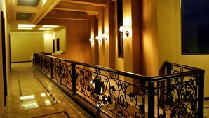Corredor de hotel com grades pretas elaboradas, arandelas nas paredes e piso de mármore.