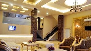 Um hall de entrada luxuoso, com colunas de mármore, mobiliário ornamentado, uma escadaria imponente e um candelabro.
