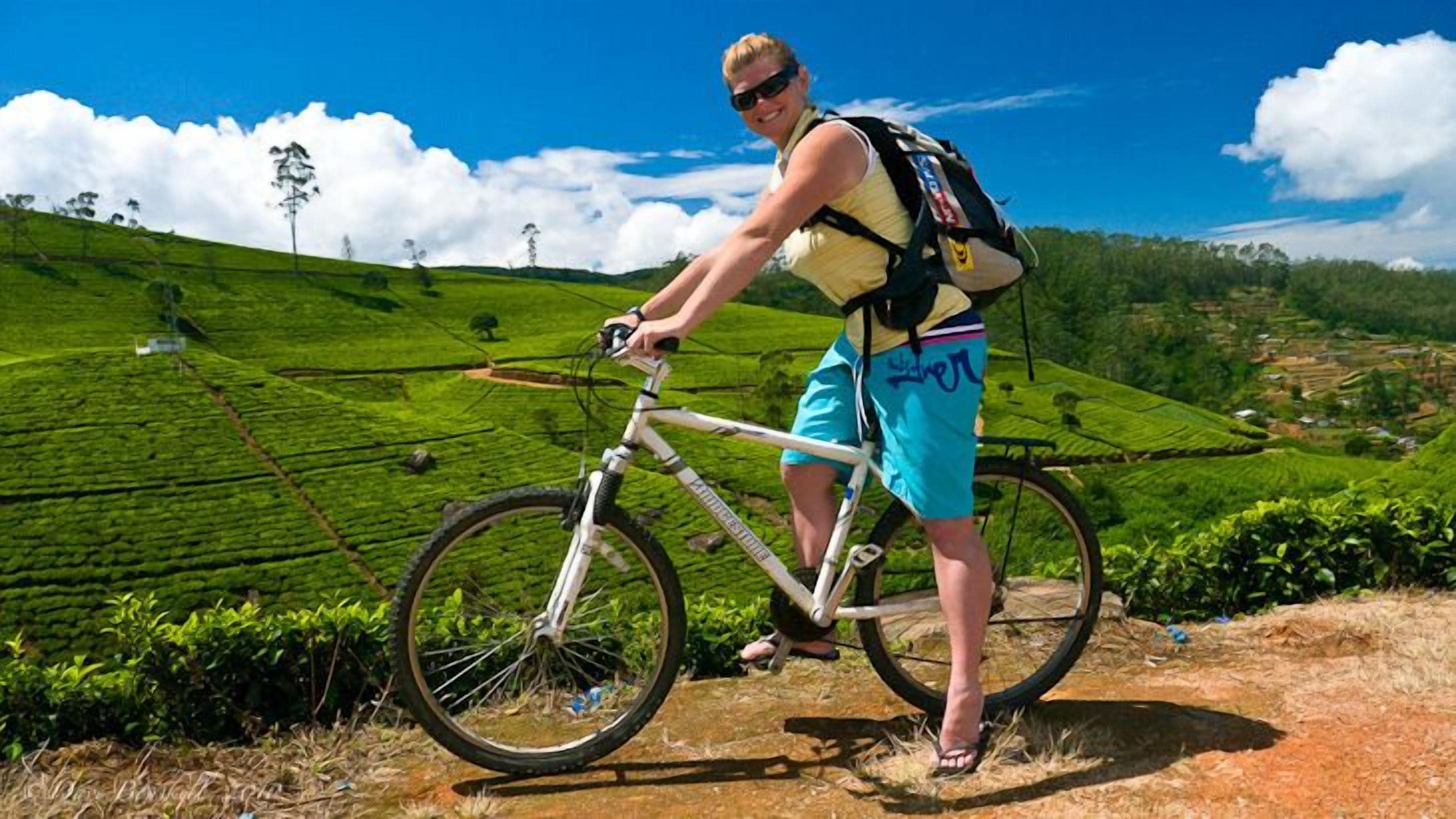 Une personne à vélo, portant un sac à dos, se tient sur un chemin de terre, avec un paysage verdoyant en arrière-plan.