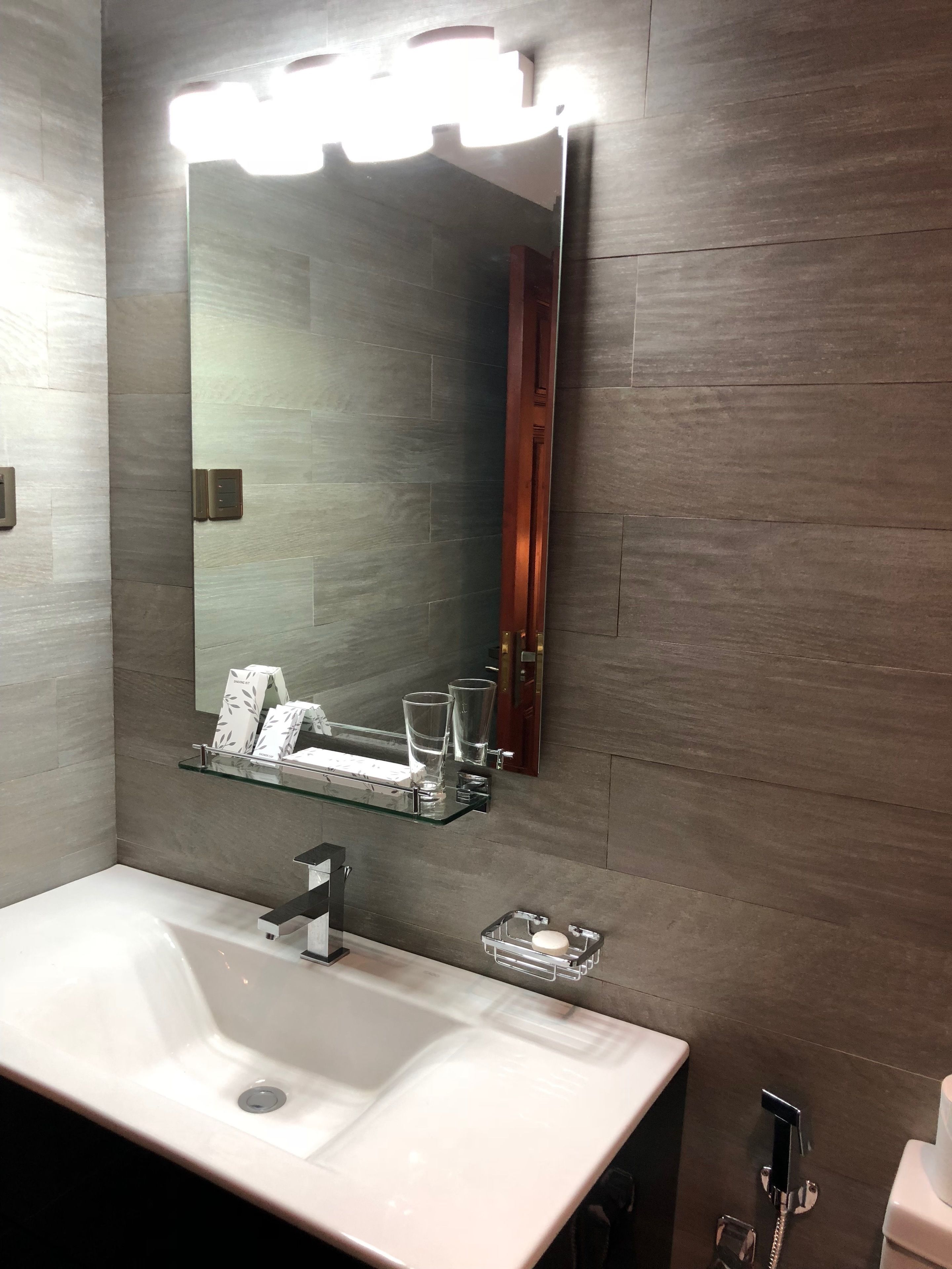 A modern bathroom with a large mirror, a white sink, and a glass shelf with toiletries.