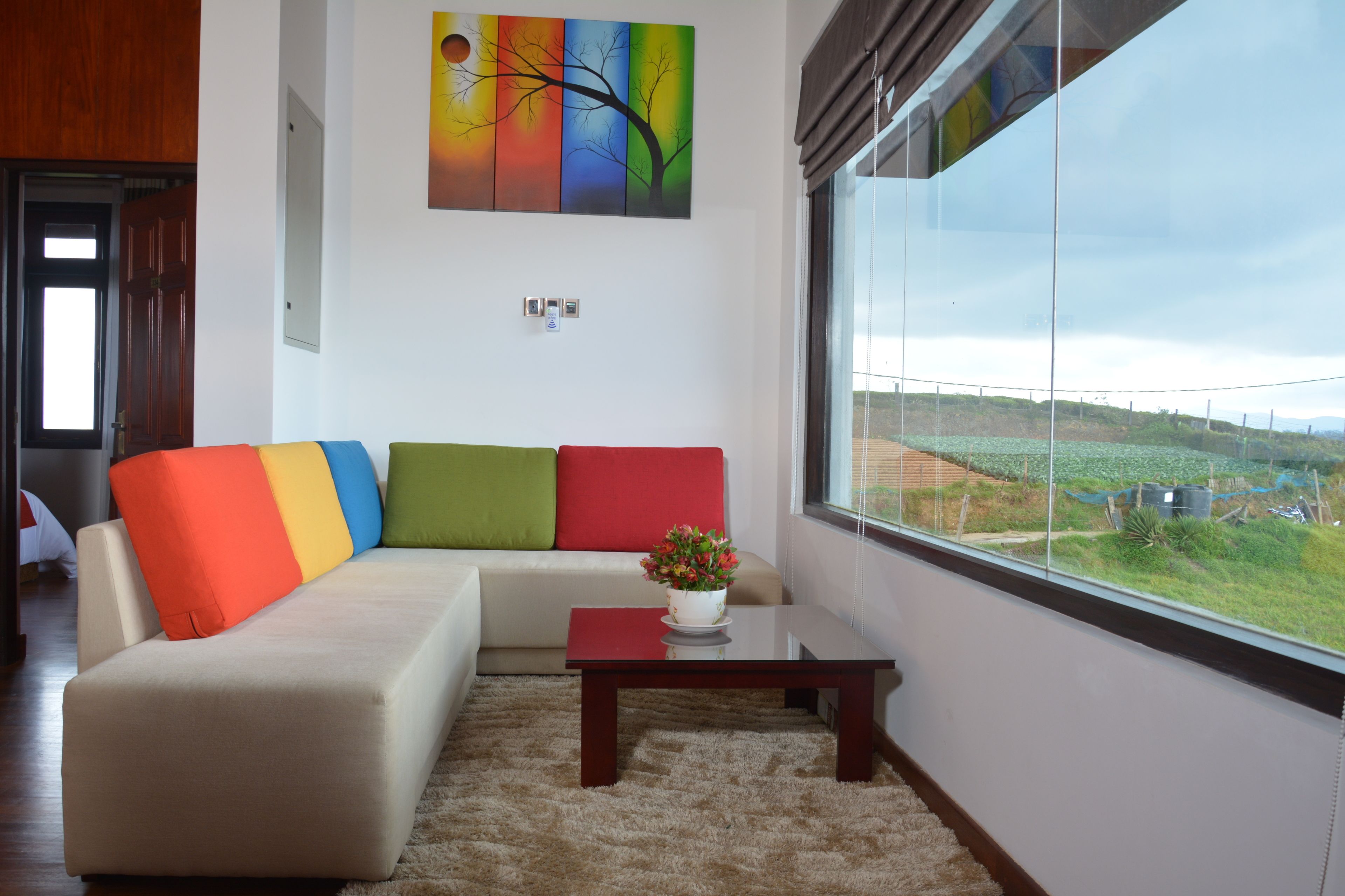 A modern living room with a beige sofa, colorful cushions, a small table with a flower, and a large window offering a view of a green landscape.