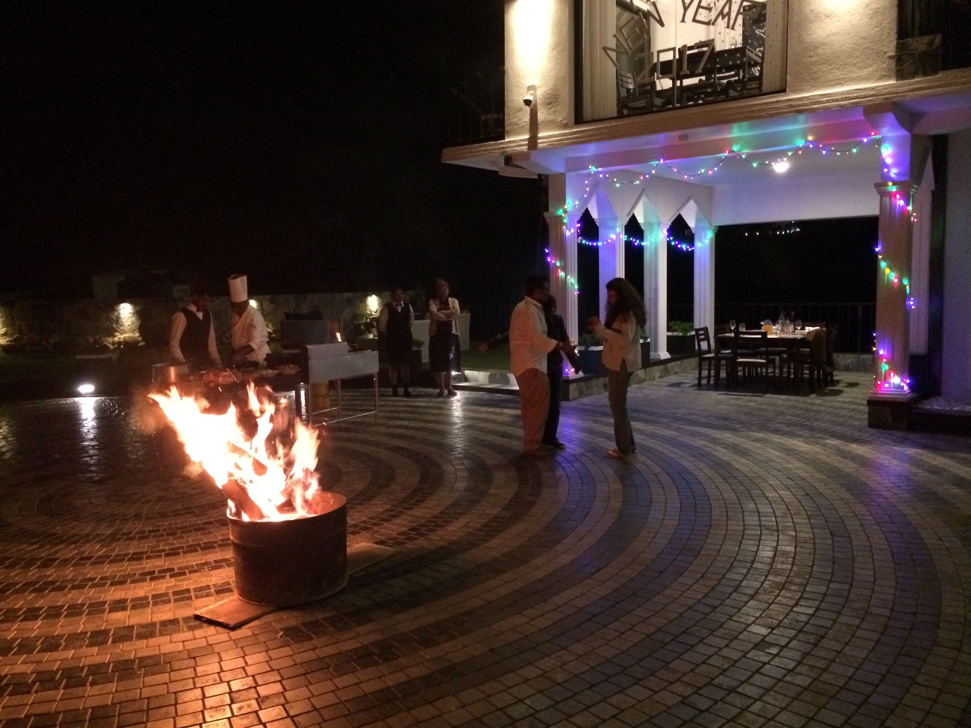 A fire pit in the center of a paved area with people around it.