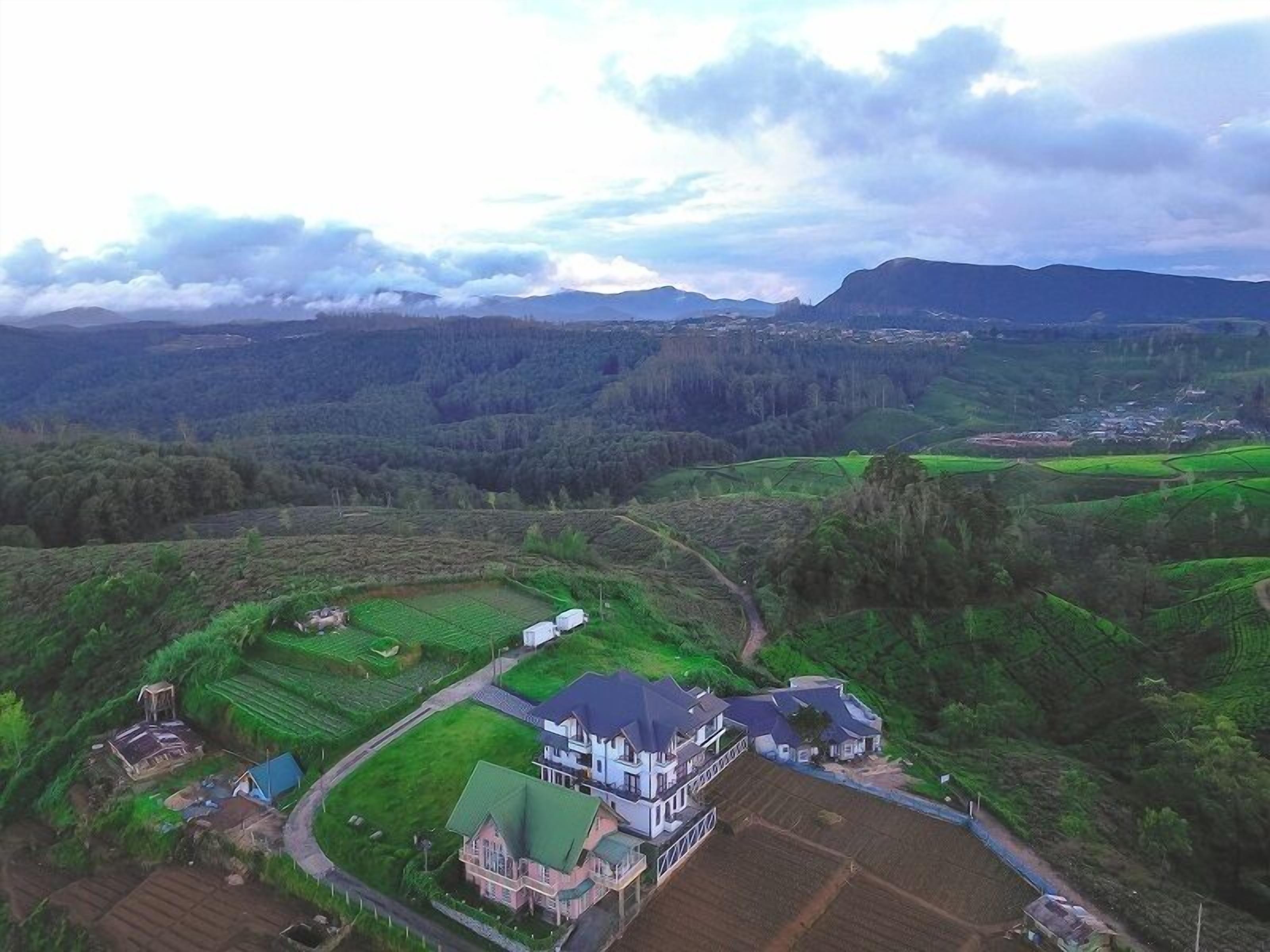 Luchtbeeld van een landelijk gebied met een groot huis, omringd door groene velden en een klein dorpje in de verte.