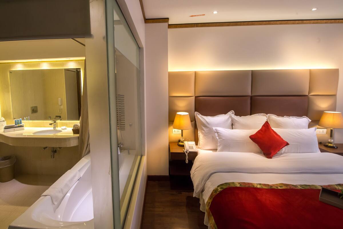 Habitación de hotel con una cama grande, lámparas de noche, una mesita de noche, un espejo y un baño con lavamanos e inodoro.