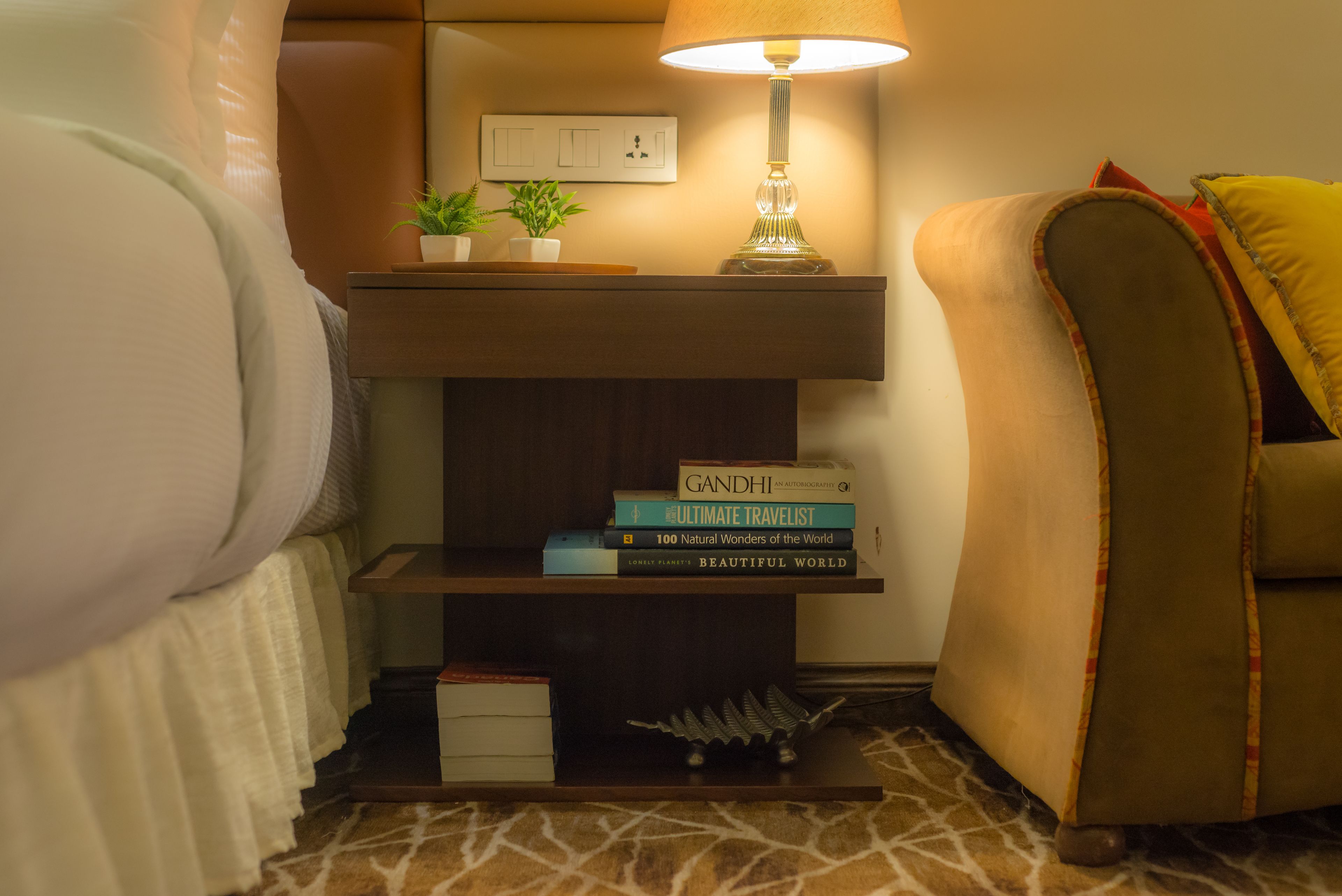 Habitación de hotel ordenada con una cama, una lámpara de noche, un pequeño estante con libros y una planta, además de un sofá con cojines.