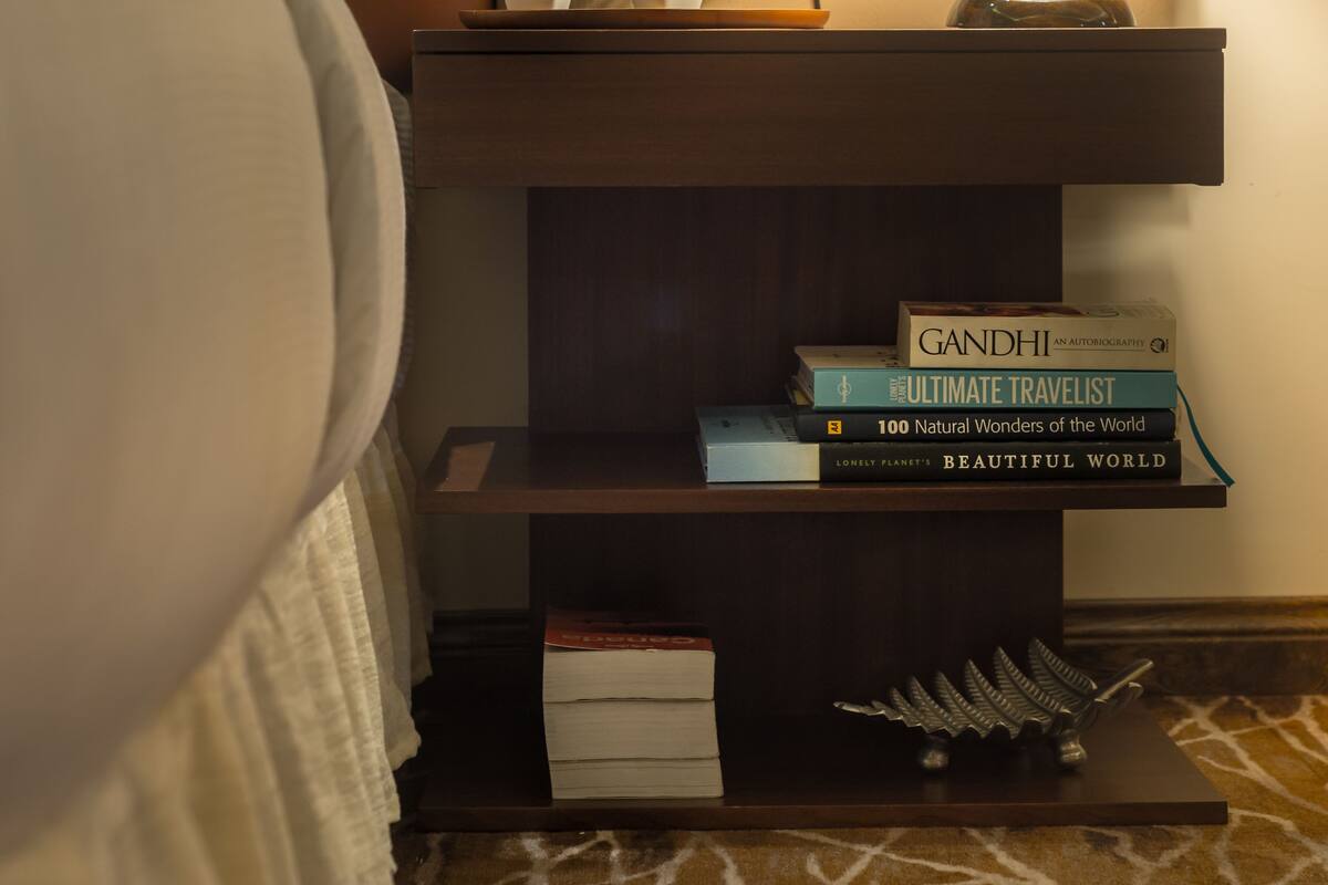 Una habitación de hotel ordenada con cama, lámpara de noche, mesita de noche con libros y una planta pequeña.