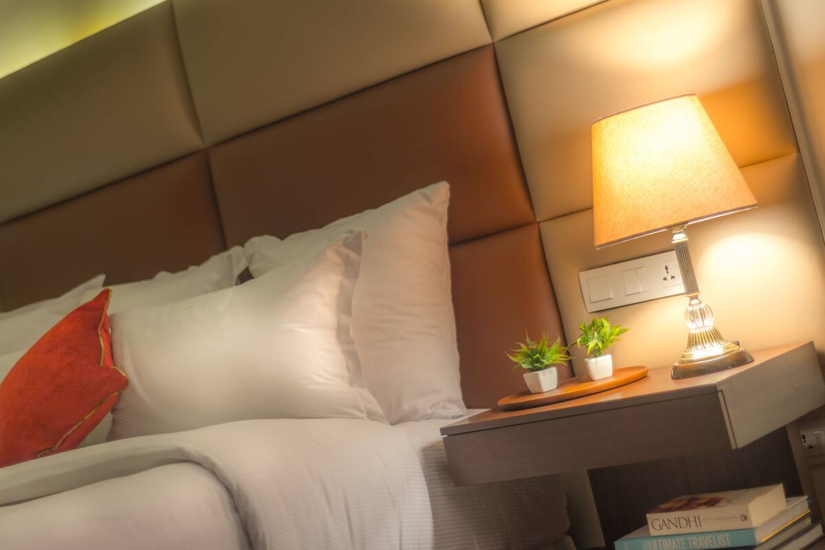 Una cama bien hecha con ropa blanca y una almohada roja, una mesita de noche con lámpara, dos pequeñas macetas con plantas y una pila de libros.