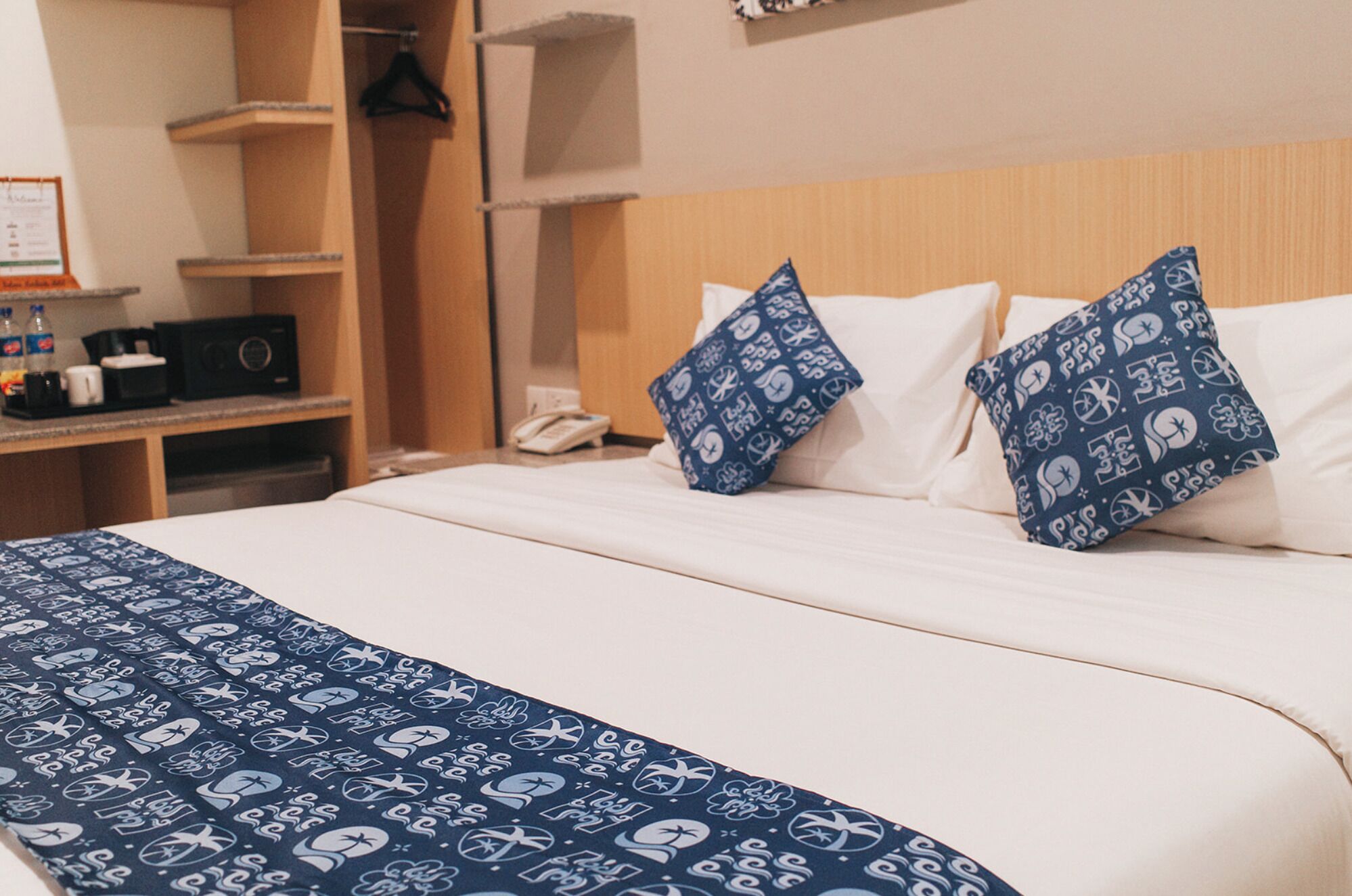 A neatly made bed with white sheets and two blue pillows with white patterns, a wooden headboard, and a bedside table with a radio and a phone.