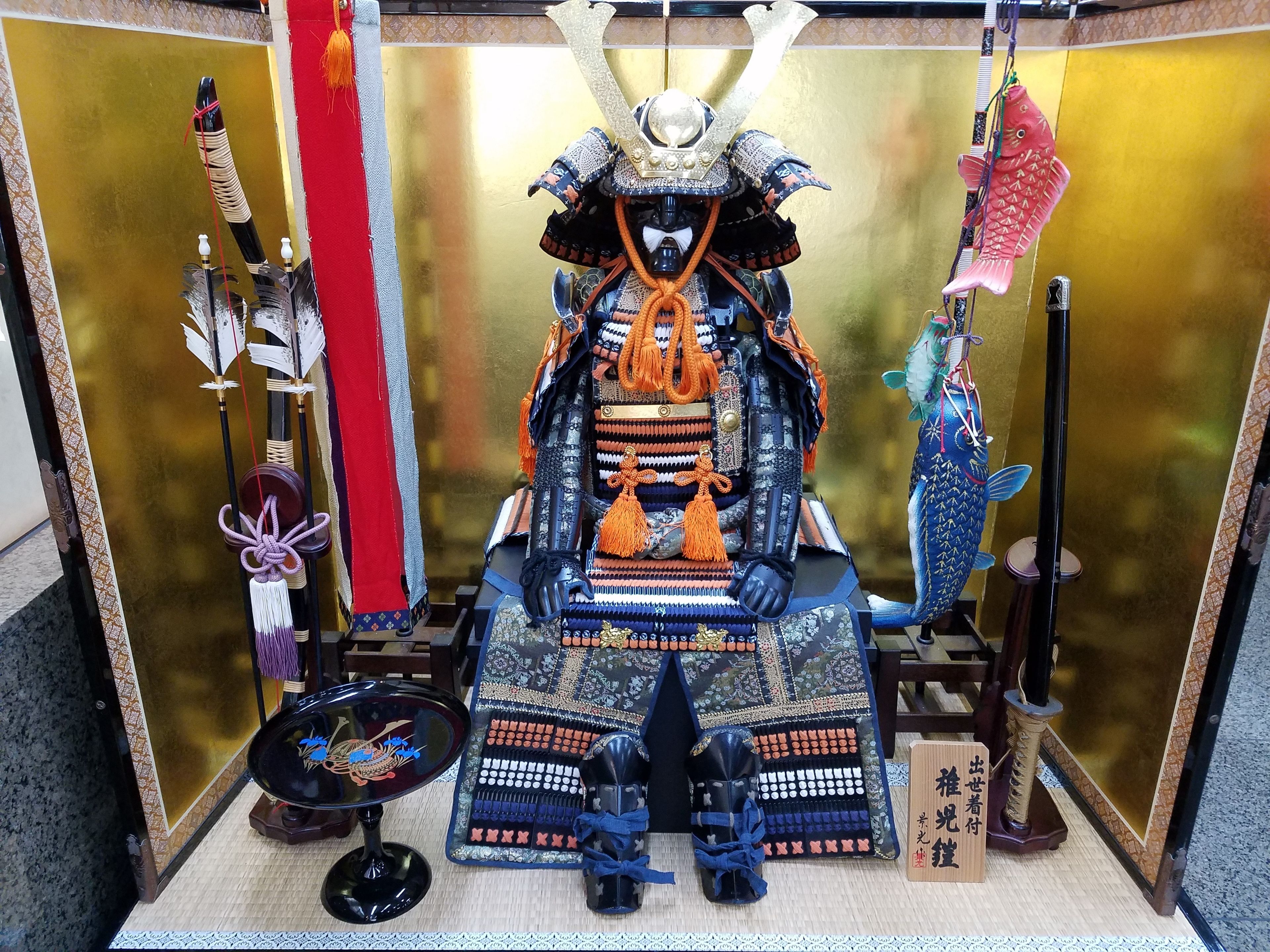 A display of a samurai armor set with a sword, a fish-shaped ornament, and a traditional Japanese fan.