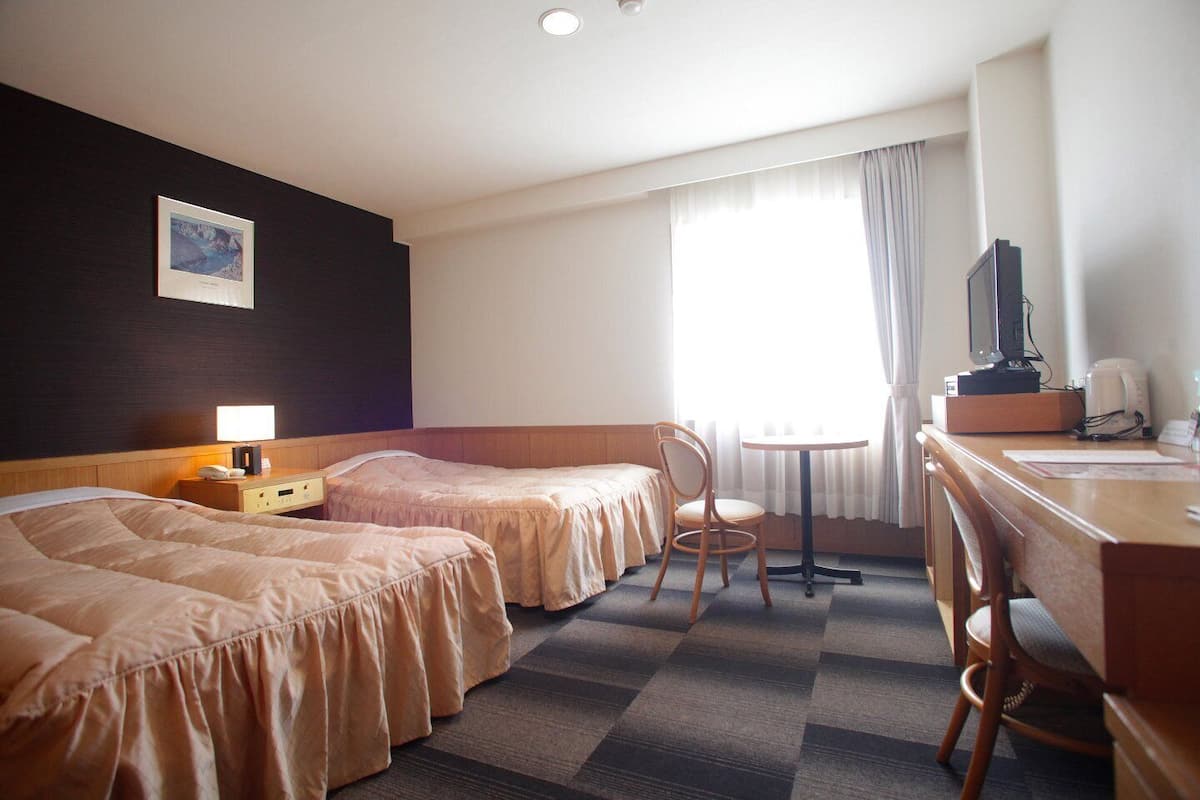A hotel room with two beds, a desk with a chair, a television, and a window with curtains.