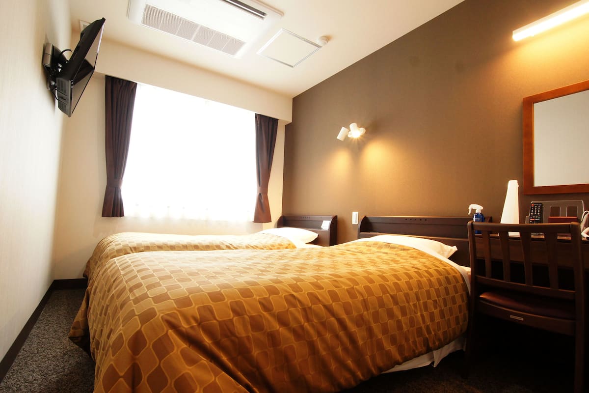 Une chambre d’hôtel avec deux lits, un bureau, une chaise, un miroir et une fenêtre avec des rideaux.