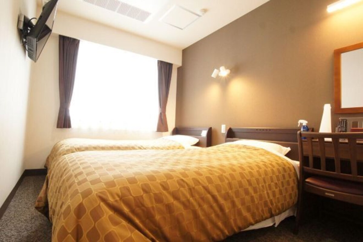 Quarto de hotel com duas camas, uma televisão na parede, uma mesa de trabalho com uma cadeira e uma janela com cortinas.