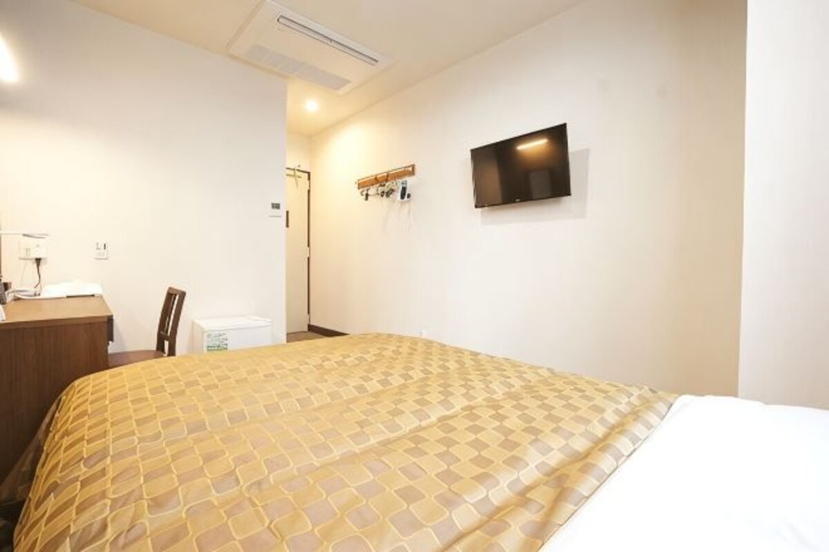 Quarto de hotel com cama, escrivaninha, cadeira, televisão na parede e porta.