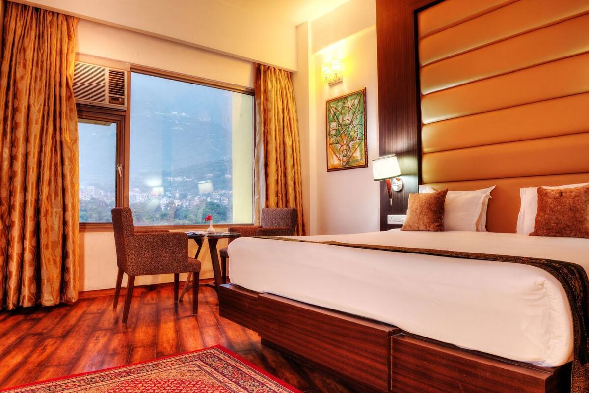 A hotel room with a large bed, a small table with chairs, a window with curtains, and a city view.
