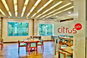 A restaurant interior with wooden tables and chairs, large windows with a view of greenery, and a ceiling with linear lighting. - Lemon Tree Hotel, Katra (Reasi)