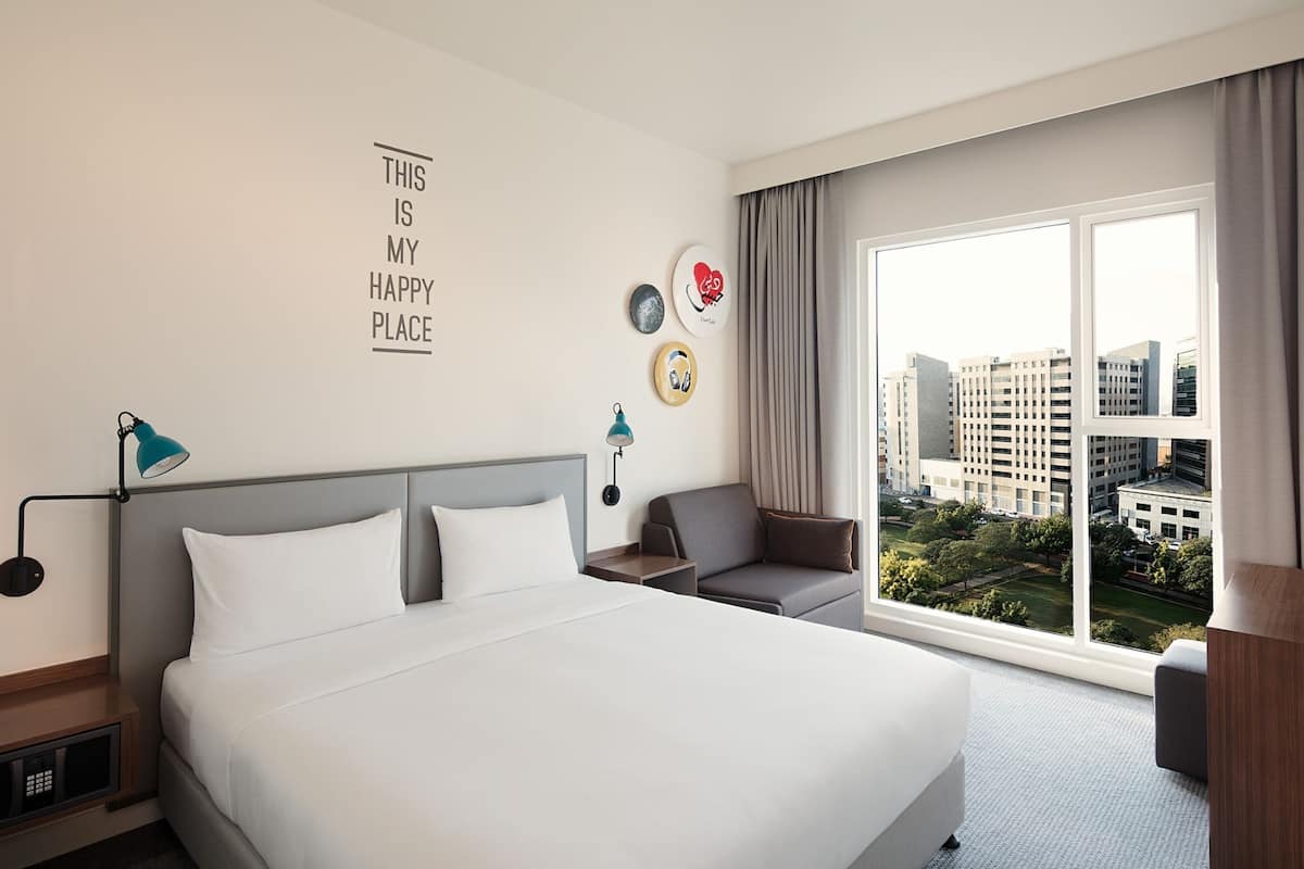 A hotel room with a large bed, a desk, a chair, and a view of the city through the window.