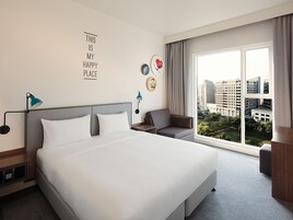 A hotel room with a large bed, a desk, a chair, and a view of the city through the window.