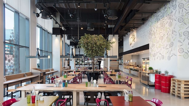 Une cafétéria spacieuse, dotée de nombreuses tables et chaises, avec un grand arbre au centre et un mur décoré d’illustrations sur le thème de la nourriture.