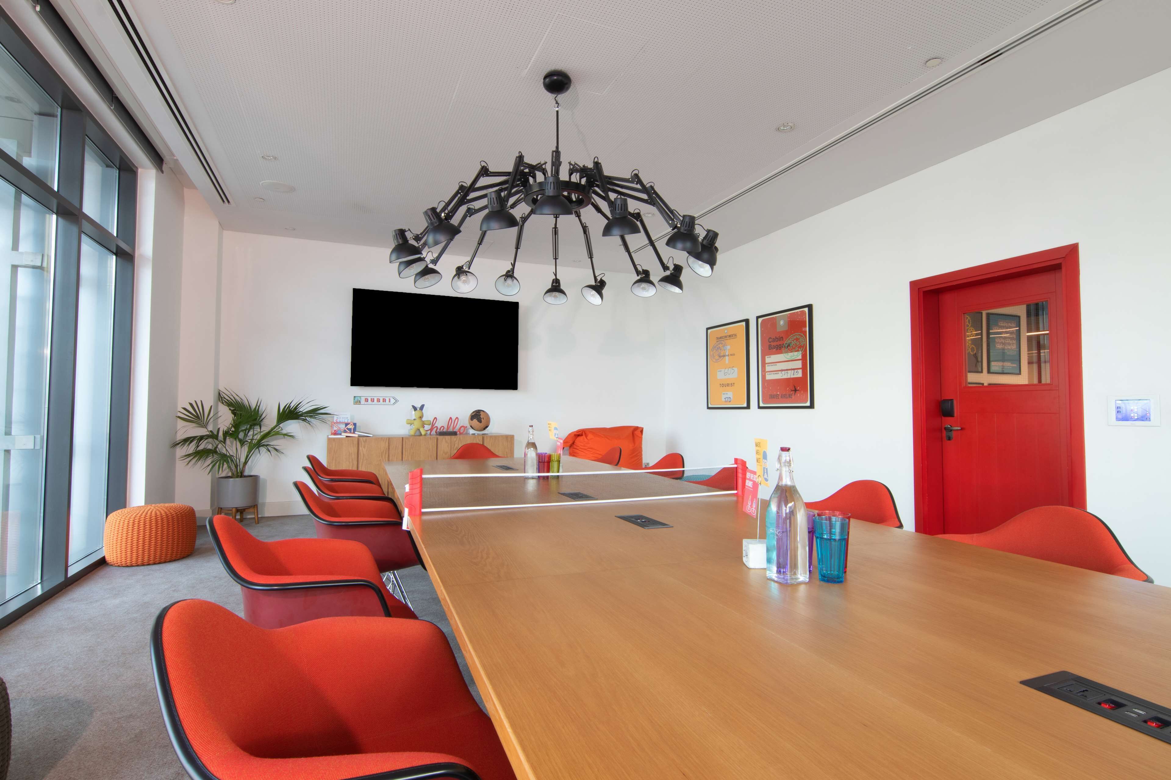 Uma sala de conferências com uma longa mesa de madeira, cadeiras vermelhas, um grande lustre, uma TV na parede e uma porta vermelha.