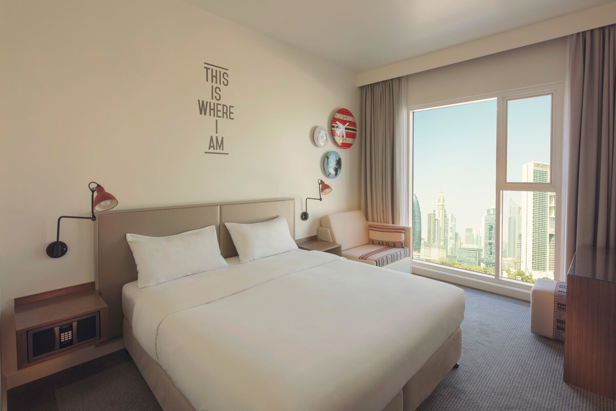 A hotel room with a large bed, a nightstand, a lamp, a chair, a window with a city view, and wall decor.