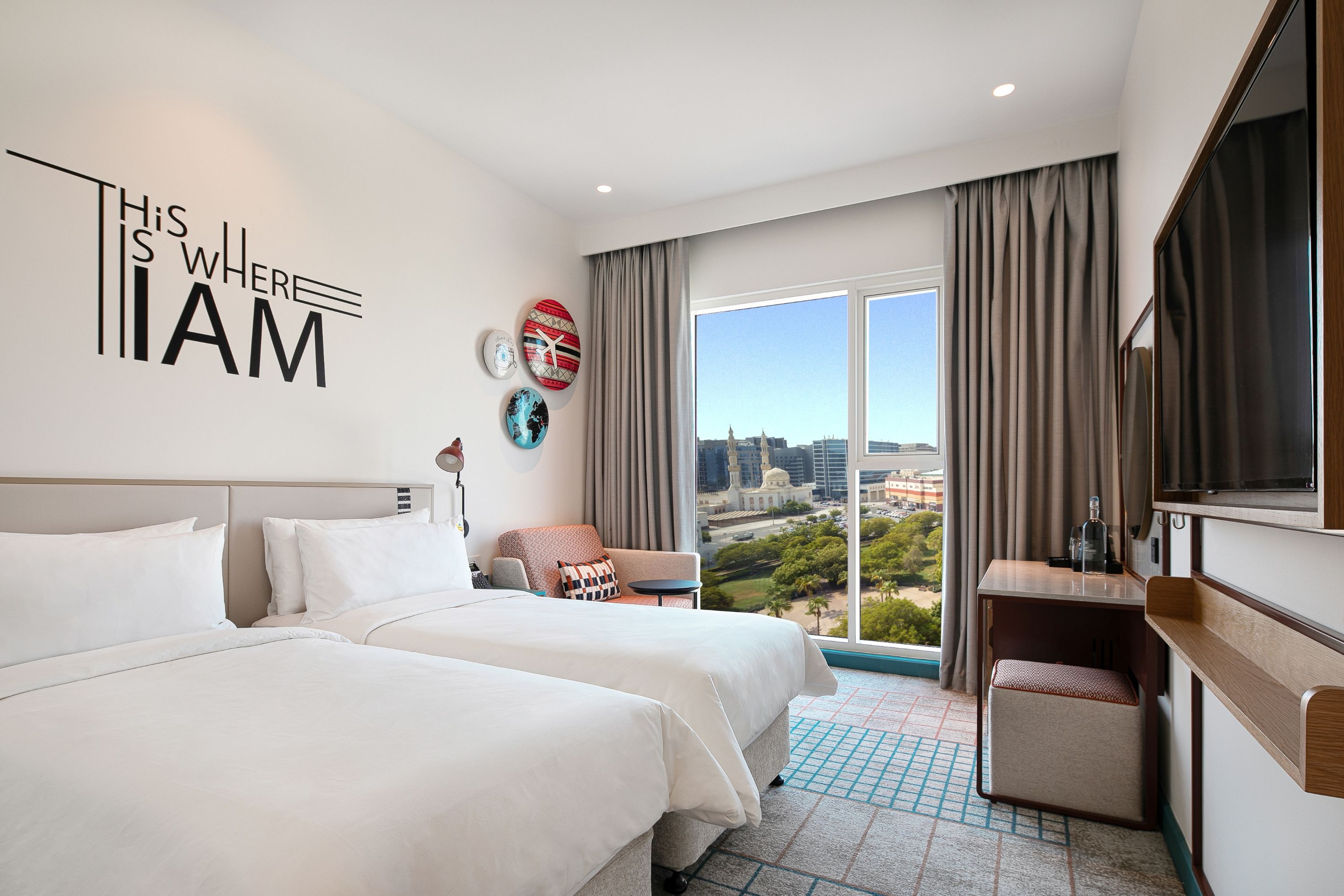 Ein Hotelzimmer mit zwei Betten, einem Schreibtisch, einem Stuhl, einem großen Fenster mit Blick auf die Stadt und einer Wand mit einem Schild.