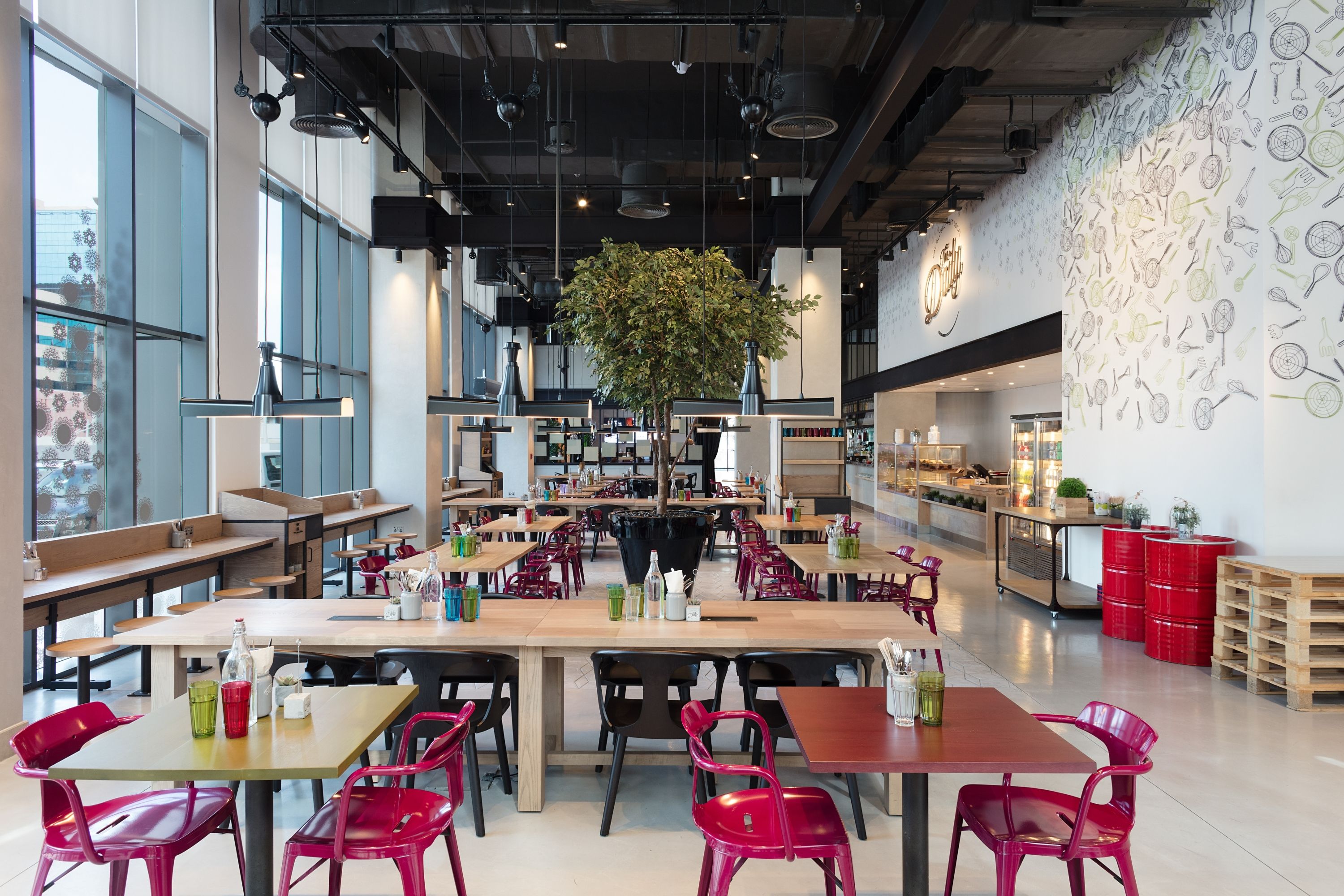 L’intérieur d’un café moderne, avec de nombreuses tables et chaises, une grande plante suspendue et un mur orné d’illustrations d’ustensiles de cuisine.