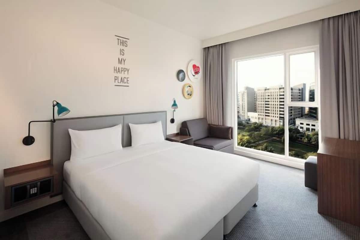A hotel room with a large bed, a desk, a chair, a window with a city view, and a wall with a motivational quote.