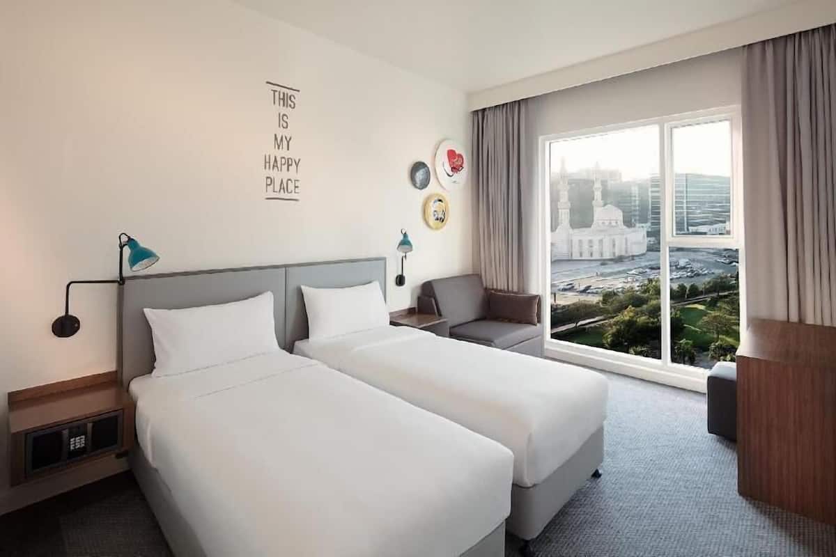 A hotel room with two beds, a sofa, a large window with a city view, and a wall with a motivational quote.