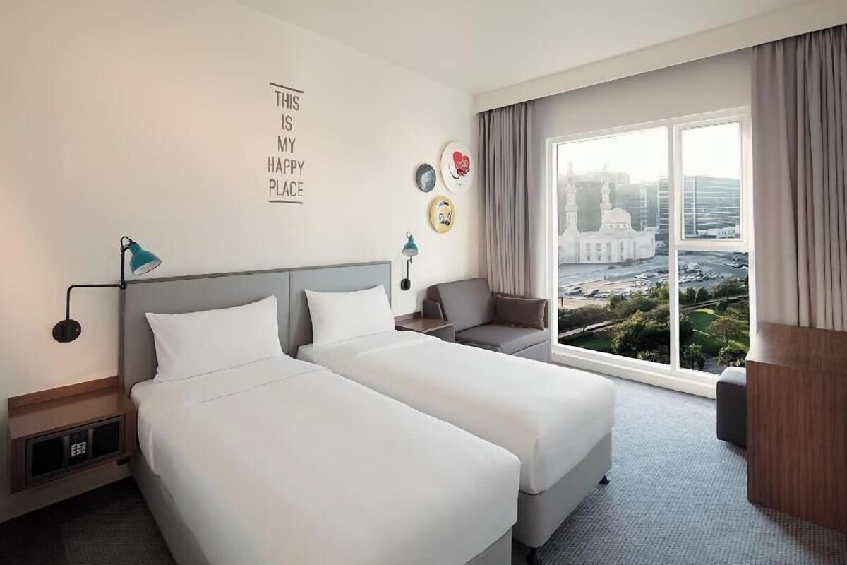A hotel room with two beds, a sofa, a large window with a city view, and a wall with a motivational quote.
