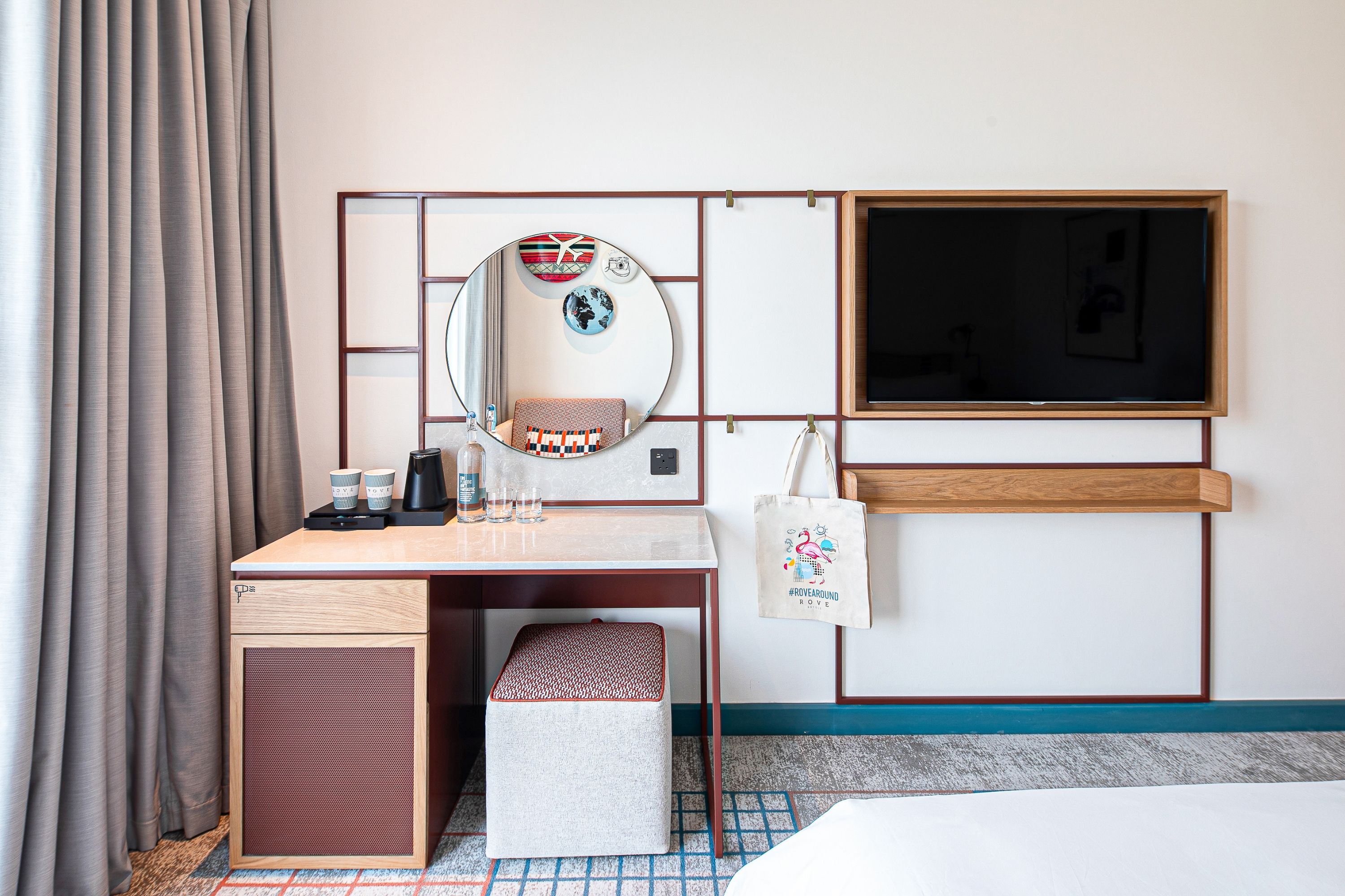A hotel room with a bed, a desk with a chair, a small stool, a mirror, a television, and a wall-mounted shelf.