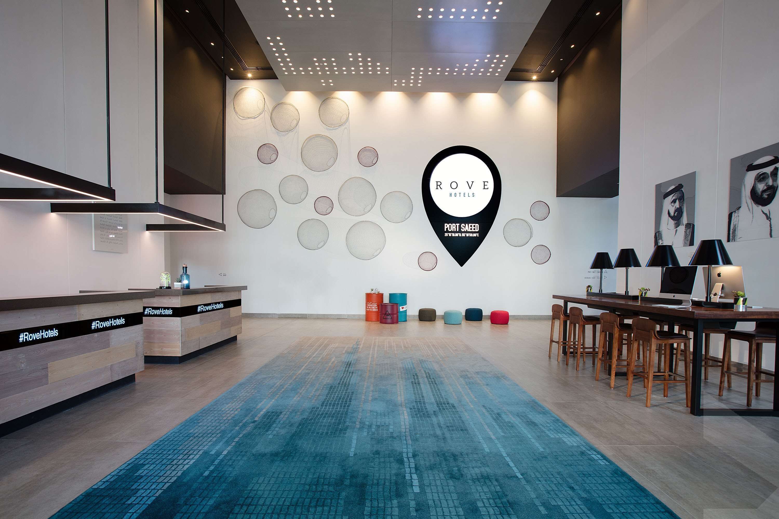 A modern hotel reception area with a large wall-mounted sign, a reception desk, and a seating area with tables and chairs.