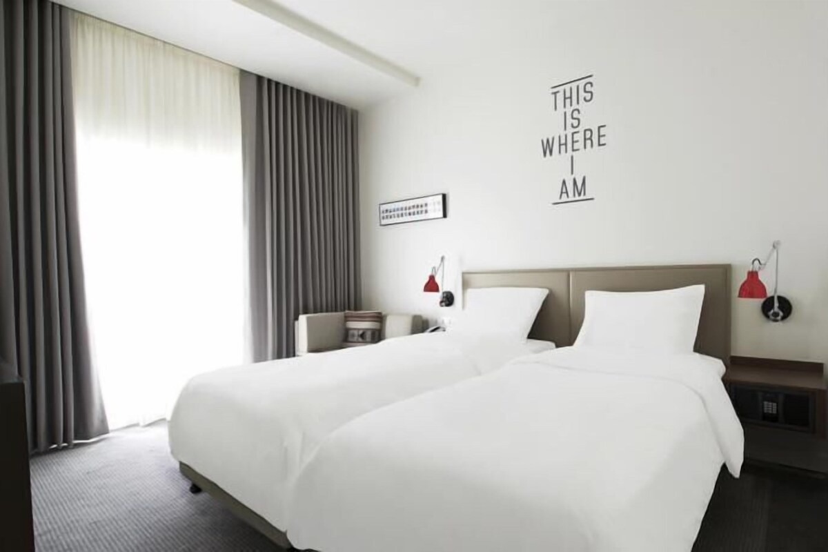 A hotel room with two beds, a large window with curtains, a wall-mounted sign, and a bedside table with a lamp.