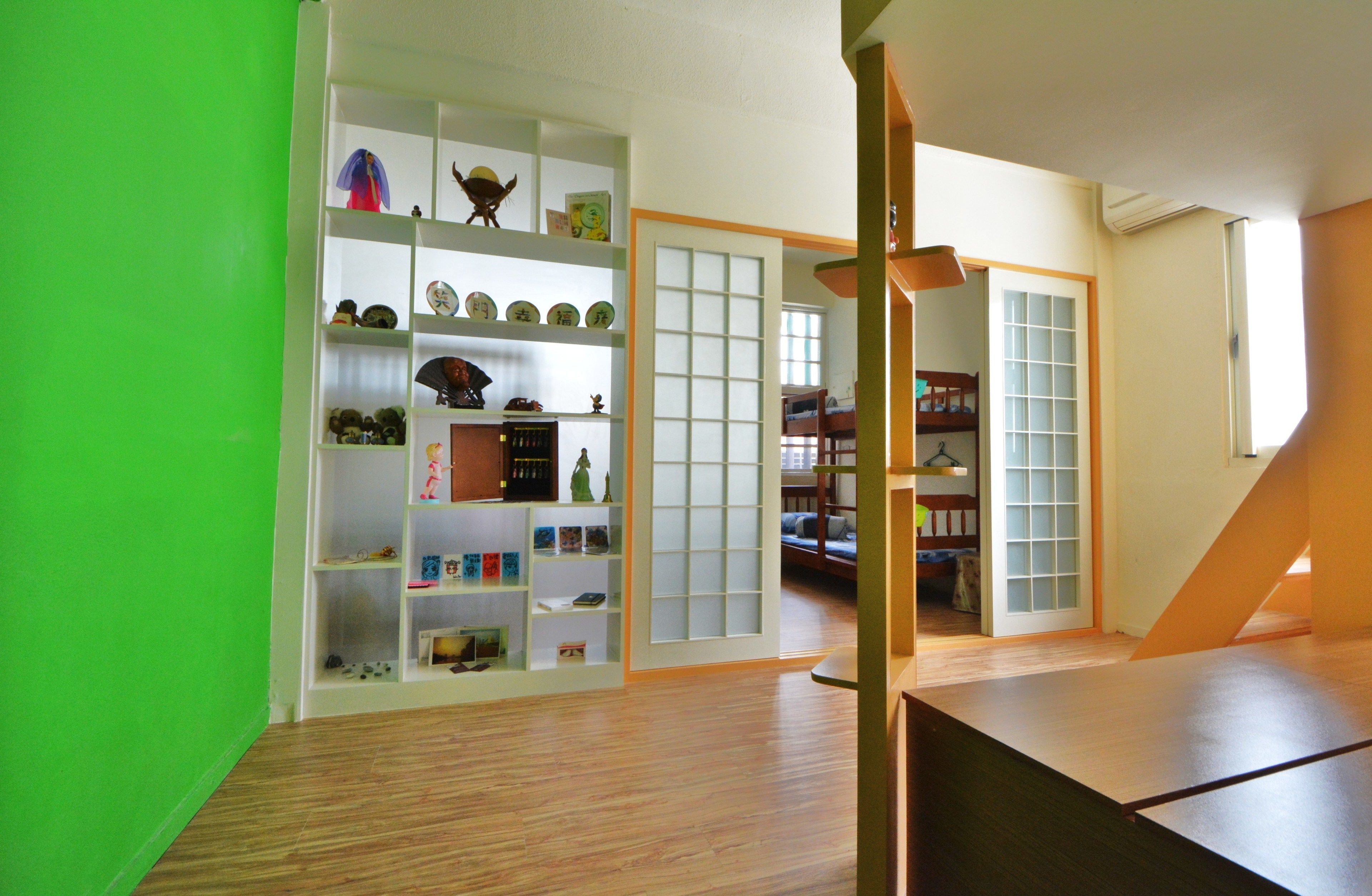 Una habitación con suelo de madera, estanterías blancas con diversos objetos, y una puerta corredera que da a otra habitación.