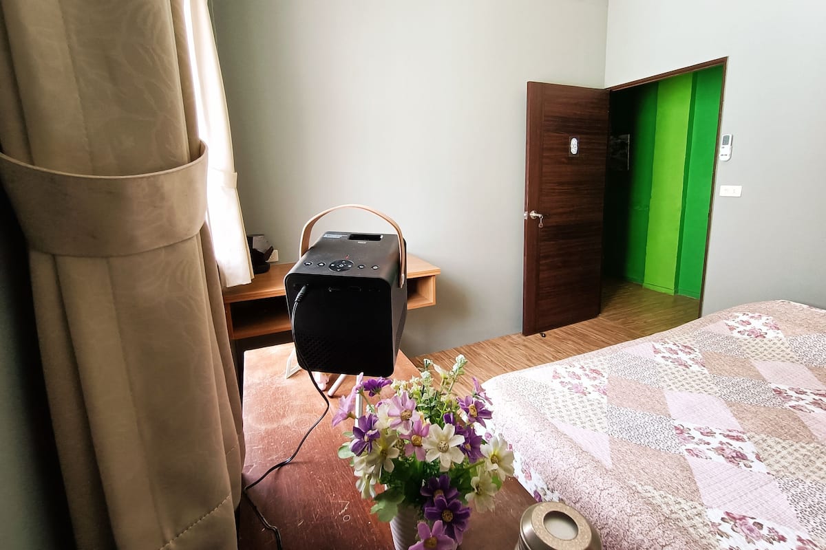 Un dormitorio con cama, escritorio, silla, una mesita, un florero con flores y una puerta que da a una habitación verde.