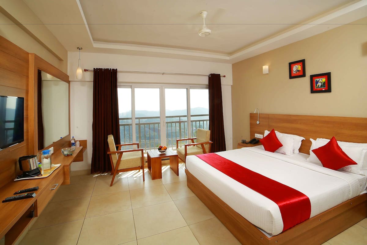 Habitación de hotel con una cama grande, un escritorio con televisor, una silla, una mesita con un frutero, y vistas a las montañas a través de las ventanas.