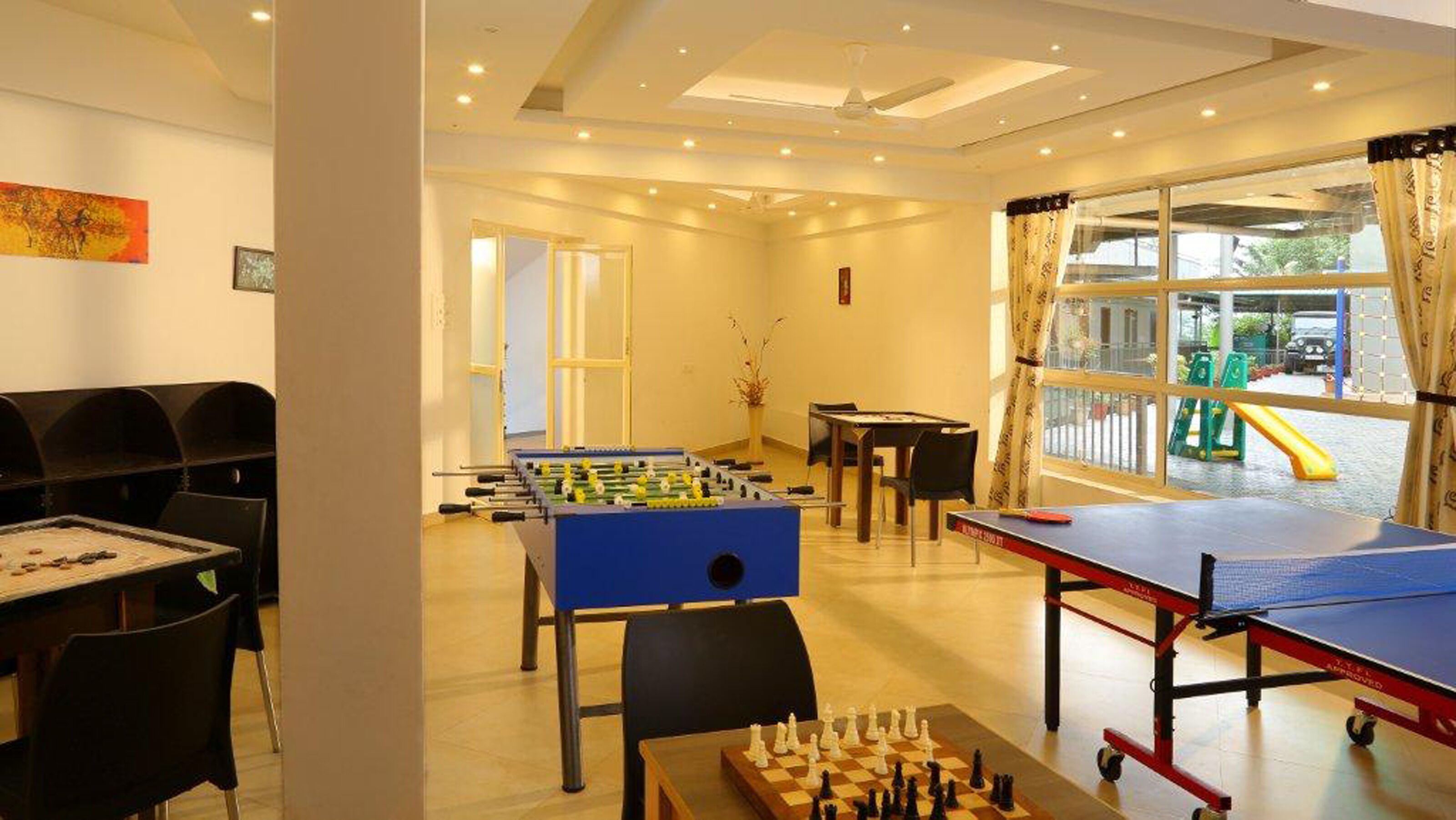 A room with a foosball table, a ping pong table, and a chess table. There is a window with a view of a playground outside.