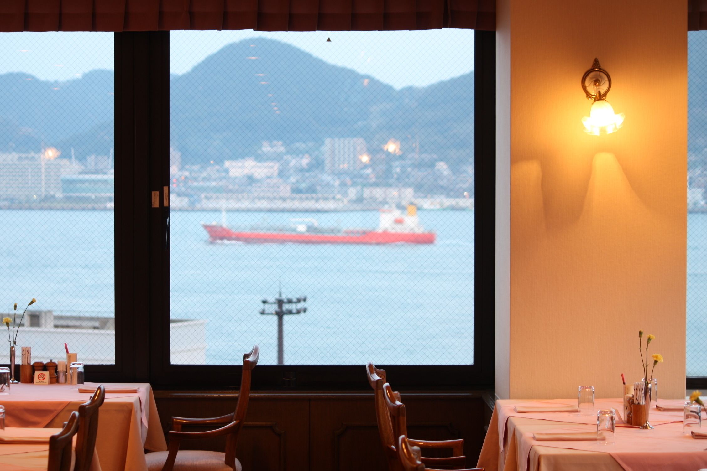 A restaurant with a table set for dining, a view of a body of water with a ship, and a wall-mounted light fixture.