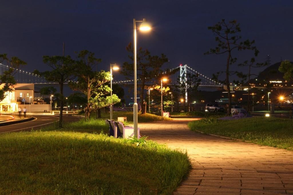A nighttime outdoor area with a paved walkway, street lamps, and a bridge in the background.