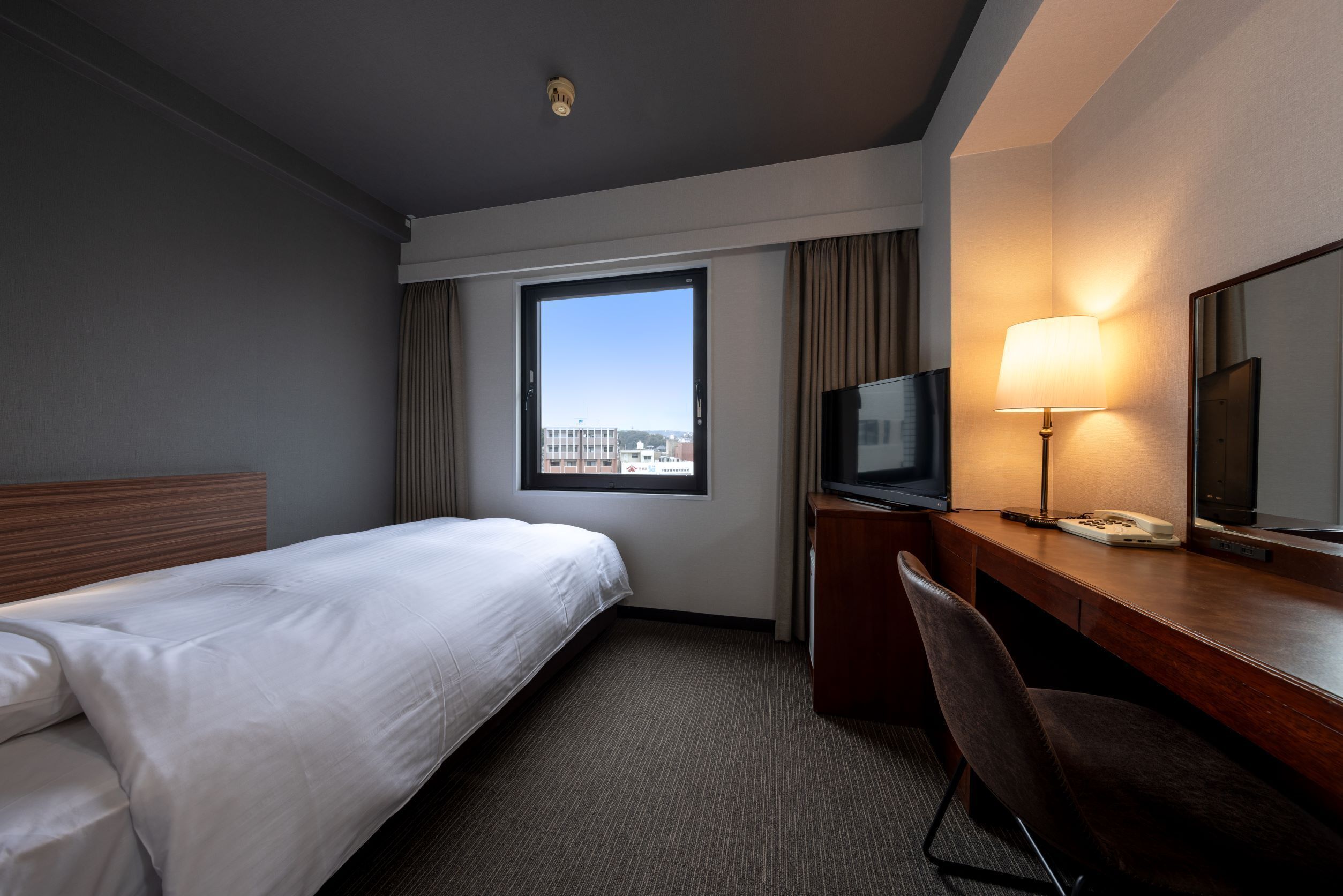 A hotel room with a bed, a desk with a lamp, a TV, and a large window.