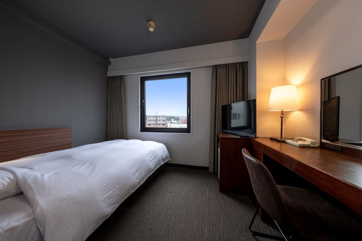 A hotel room with a bed, a desk with a lamp, a TV, and a large window.