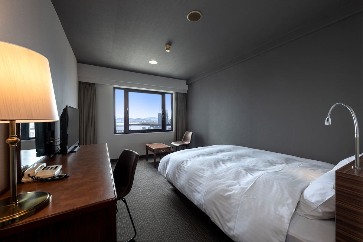 A hotel room with a bed, desk, chair, lamp, and a view of the city through large windows.